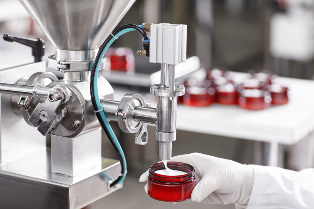 Hard mechanism of packing ready beauty cream into red container. A factory worker dressed in white lab gown and gloves holding red container under tap pouring ready cream. Production process of cream